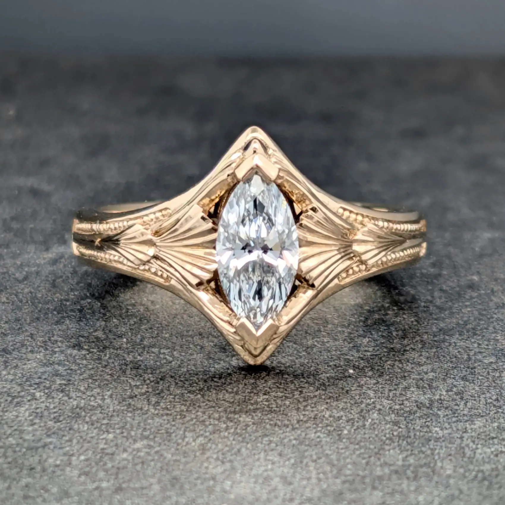 Close-up of a rose-gold ring featuring a large central diamond and carved leaf-pattern band on a dark gray surface.