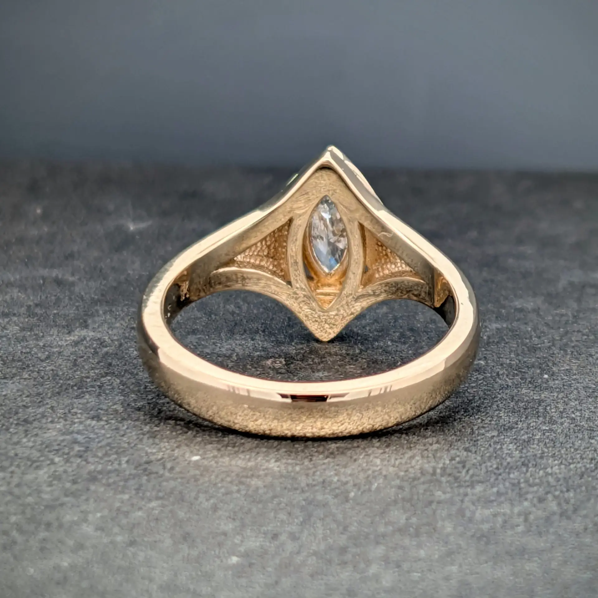 Close-up of a gold ring with a triangular, faceted setting and a central clear gemstone on a dark textured surface.