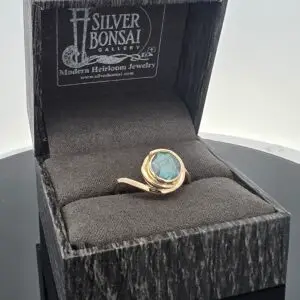 Gold ring with a blue cabochon gemstone placed in a black presentation box with Silver Bonsai Gallery branding on the lid (logo and website visible).