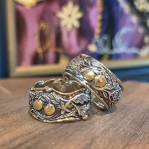 Two ornate silver bracelets with gold accents resting on a wooden surface, against a blurred colorful background of purple fabrics and a frame.